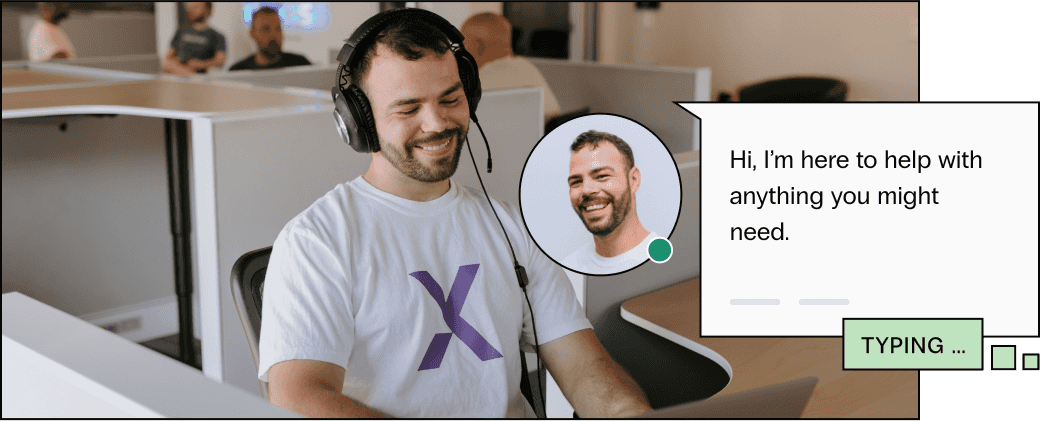 A man in a light tshirt smiles as he assists a customer via chat, his colleagues are in the background